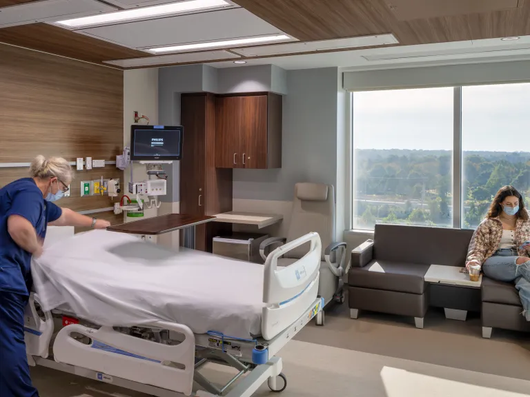 Interior day of a hospital room with a nurse making the bed and a woman sitting by the window on her phone