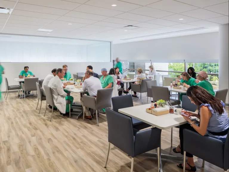 Cafeteria with many people in medical uniforms eating lunch, talking, and working at tables.