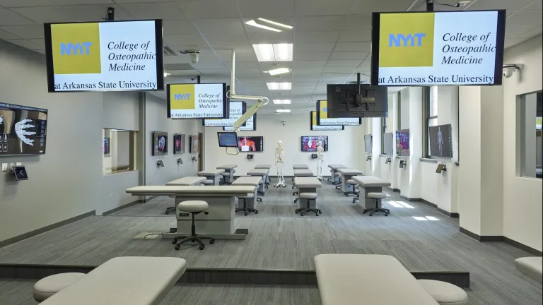 Interior of osteopathic classroom with doctor office style tables and rolling chairs, featuring multiple televisions at each station
