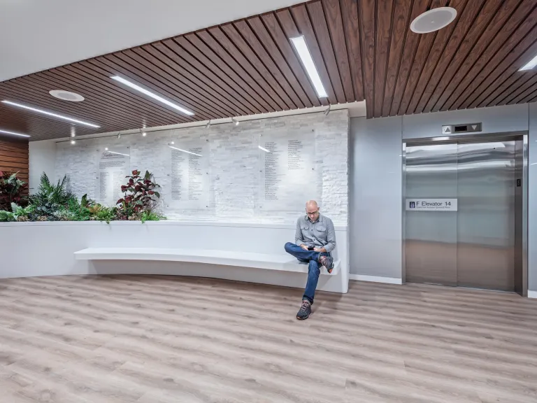 Elevator entrance of Care Center with dark wood paneled ceiling, white bench, and white tile wall with donor plaques